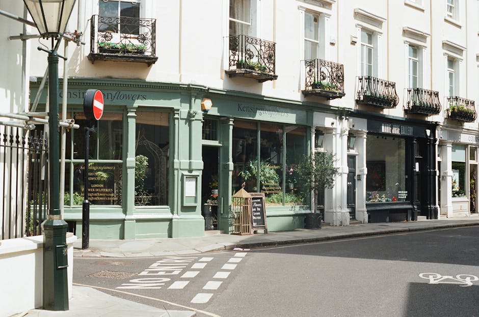 Street view of a florist shop in Knightsbridge with a mint green exterior, large glass windows displaying floral arrangements, and a blackboard sign outside. The shop features decorative iron balcony 
